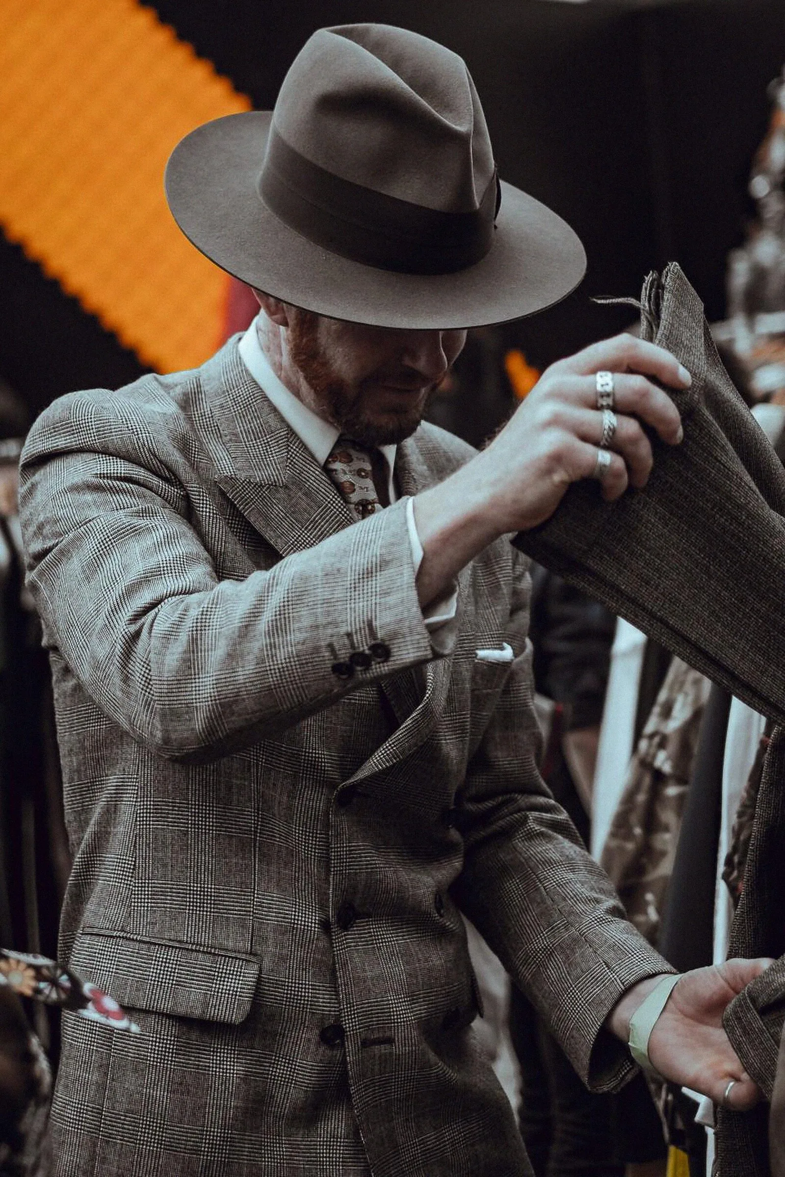 A man in a brown tweed suit and felt hat adjusting a fabric swatch.