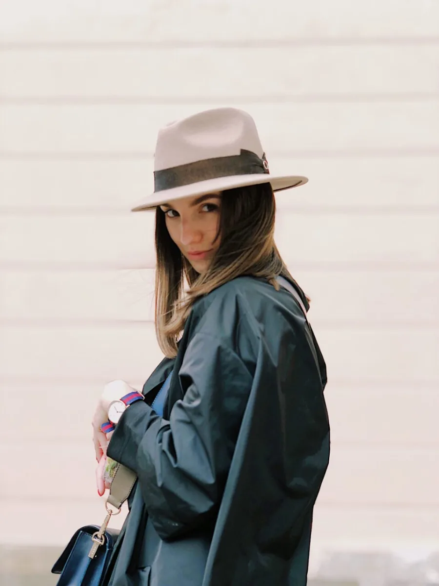 A woman in a felt hat and dark trench coat against a pale wall.