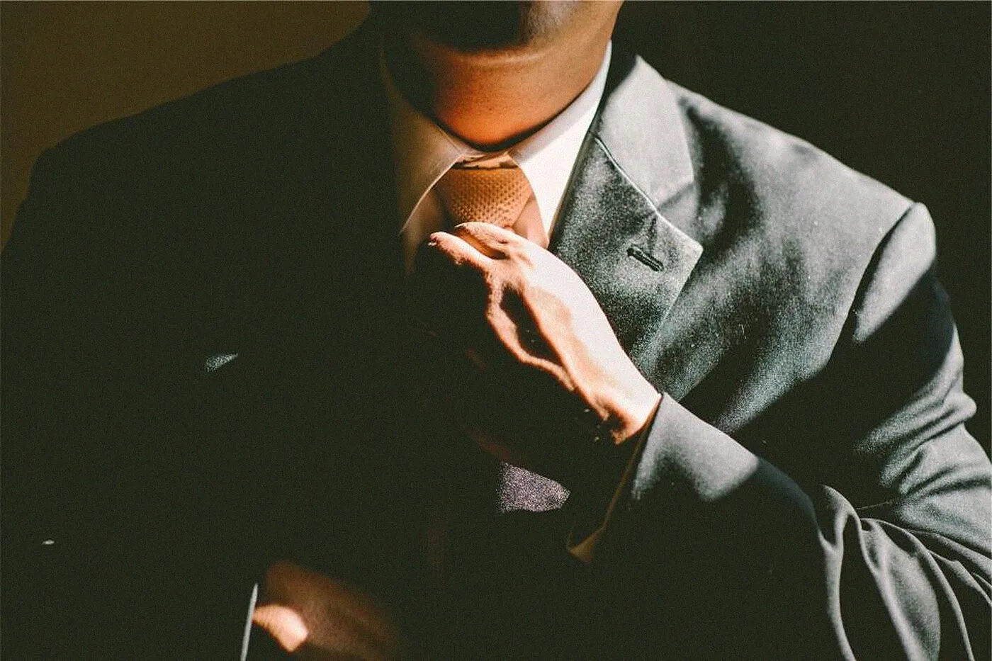 A man in a dark suit straightening his tie in low light.